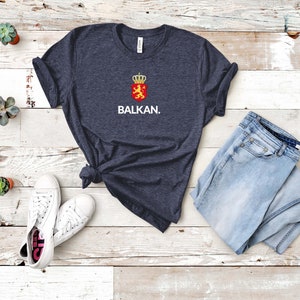 K&ouml;nnte beinhalten: Ein dunkelblaues T-Shirt mit einer wei&szlig;en Grafik eines L&ouml;wen in einem Schild mit einer Krone oben. Der Text "BALKAN." befindet sich unter der Grafik.