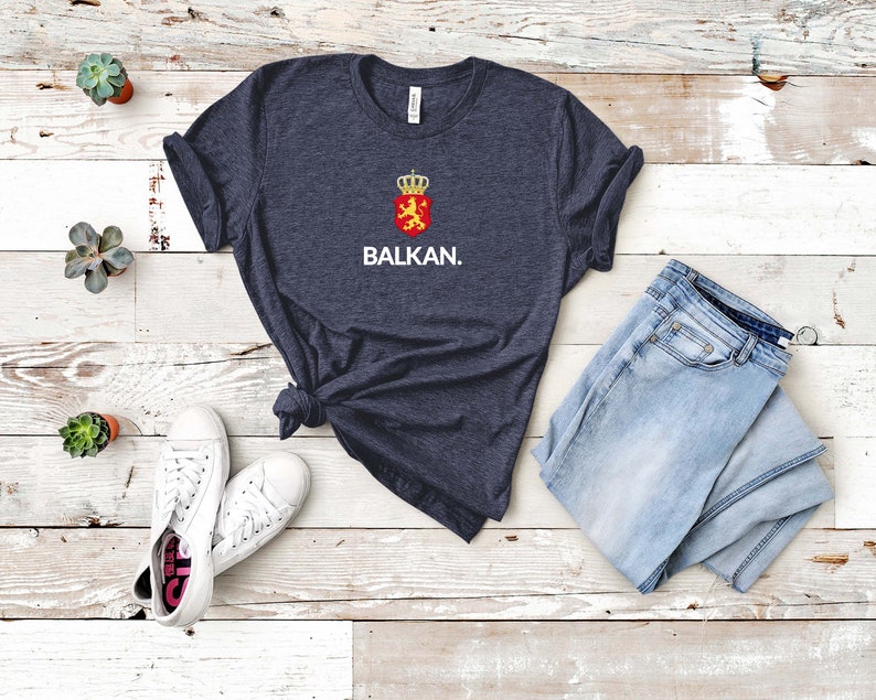 K&ouml;nnte beinhalten: Ein dunkelblaues T-Shirt mit einer wei&szlig;en Grafik eines L&ouml;wen in einem Schild mit einer Krone oben. Der Text "BALKAN." befindet sich unter der Grafik.