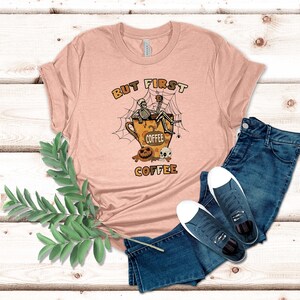 May include: A pink t-shirt with the text "But First Coffee" and a skeleton sitting in a coffee cup with a spider web and pumpkins.