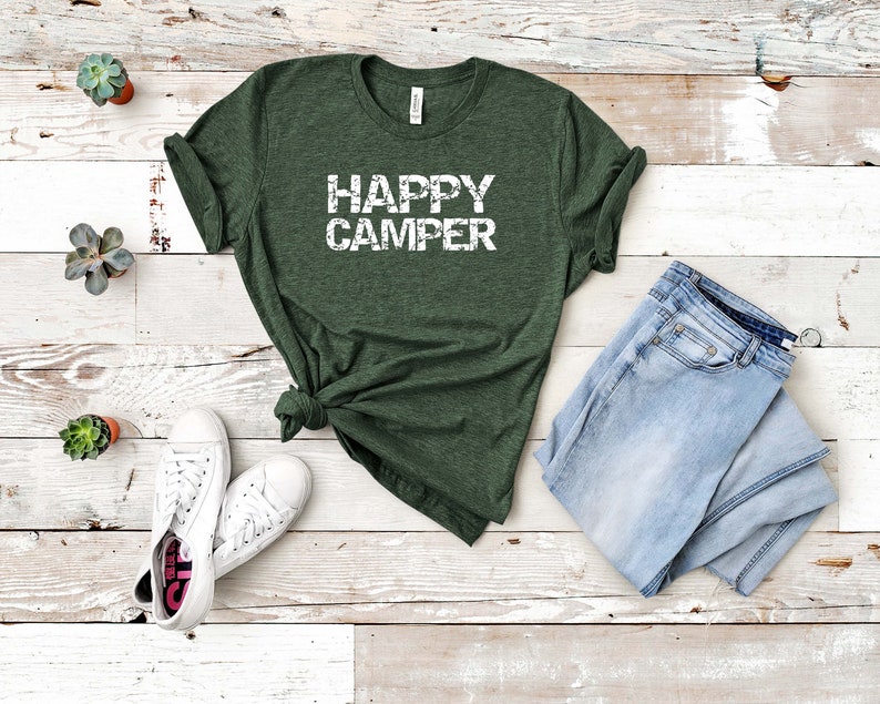 Puede incluir: Una camiseta verde con el texto "HAPPY CAMPER" en letras blancas desgastadas. La camiseta est&aacute; colocada sobre un fondo de madera blanca con un par de zapatillas blancas y unos vaqueros azules.