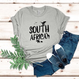 May include: A light gray t-shirt with the words "SOUTH AFRICA" in black, featuring silhouettes of birds, a giraffe, and elephants. The shirt is paired with blue jeans and navy sneakers with white soles.
