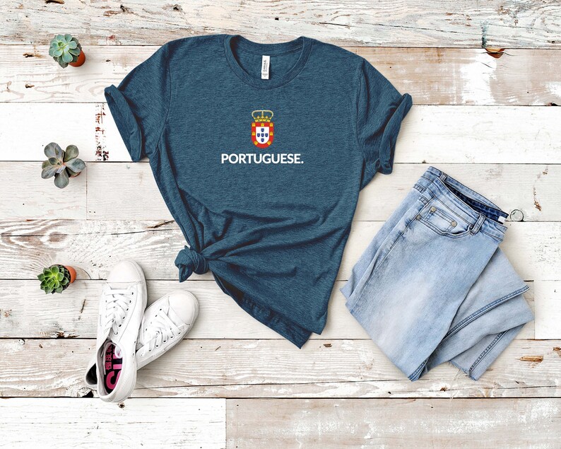 Może przedstawiać: Turkusowa koszulka z napisem "PORTUGUESE" w białym kolorze. Nad tekstem znajduje się herb z koroną i czerwono-biało-niebieskim emblematem. Koszulka jest połączona z jasnoniebieskimi dżinsami i białymi trampkami.