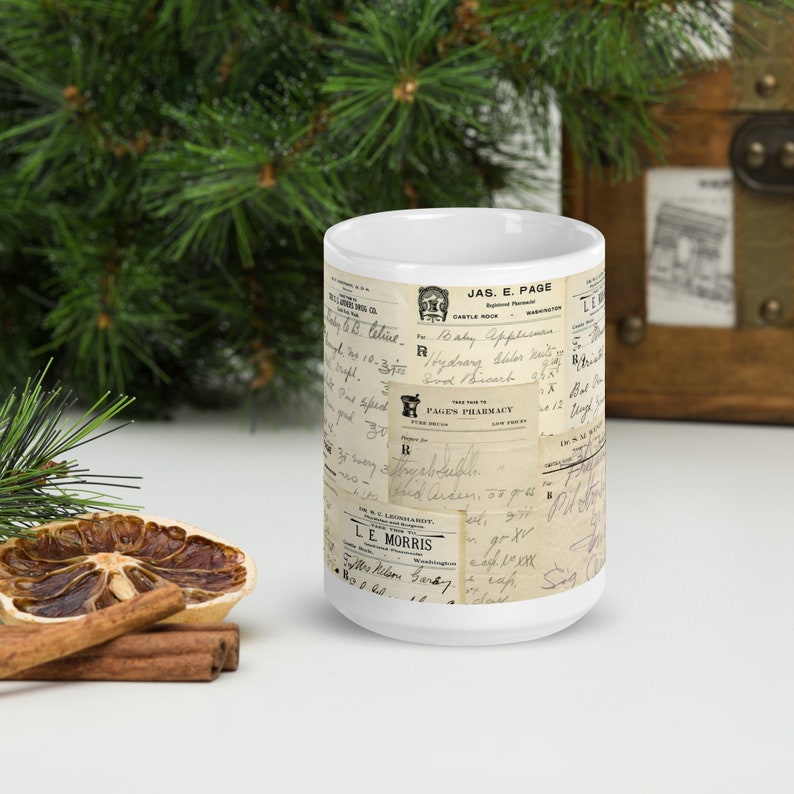 Op de afbeelding: Een witte keramische mok met een vintage ontwerp met een collage van oude handgeschreven bonnen en etiketten. De tekst op de bonnen omvat "JAS. E. PAGE", "CASTLE ROCK", "PAGES PHARMACY" en "LE MORRIS".