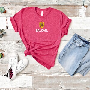 May include: A pink heather t-shirt with a white graphic of the coat of arms of Romania and the word "BALKAN." The shirt is laid flat on a white wood background with a pair of white sneakers and a pair of light blue jeans.