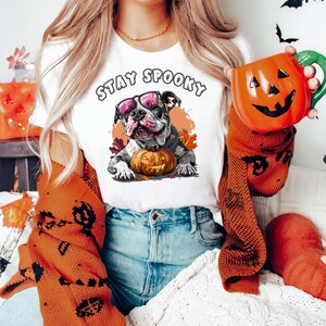 May include: White t-shirt with a black and white bulldog wearing sunglasses and holding a jack-o-lantern. The text "Stay Spooky" is printed on the shirt.