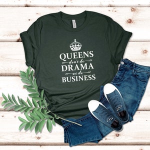 May include: A dark green short-sleeved t-shirt featuring a white graphic of a crown above the text "QUEENS don't do DRAMA we do BUSINESS," paired with blue denim jeans and a pair of dark blue canvas trainers with white laces and soles.