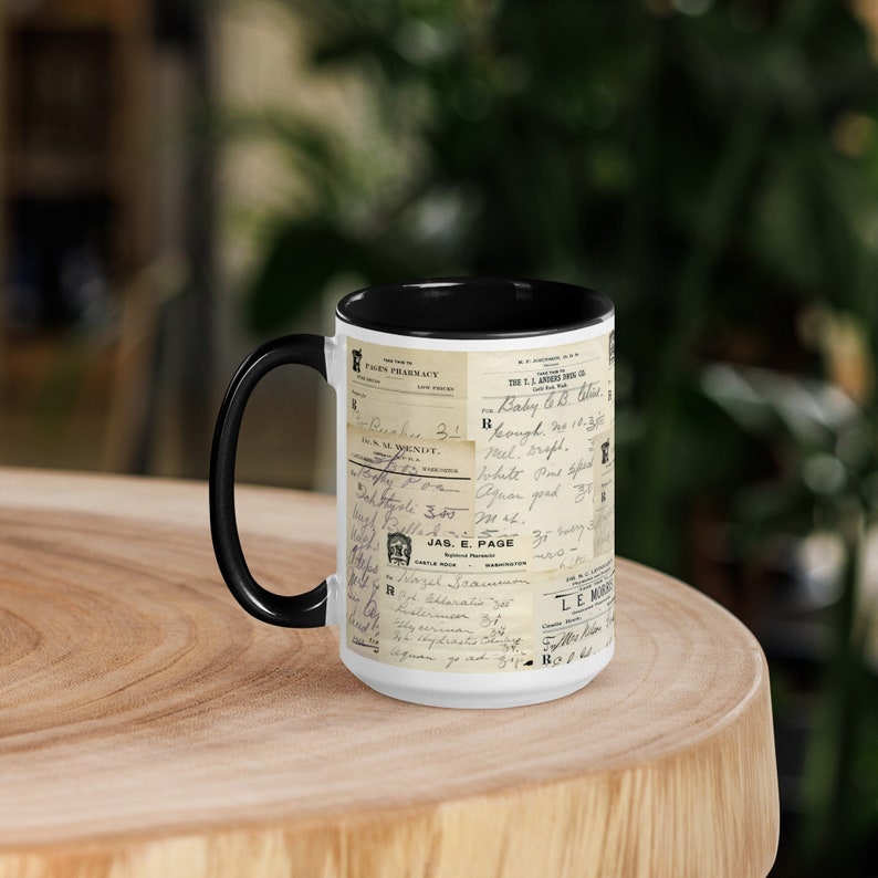 Op de afbeelding: Een witte keramische mok met een zwarte handgreep en zwarte rand. De mok heeft een vintage ontwerp met een collage van oude receptetiketten en handschrift. De tekst bevat "Baby & Cough" en "JAS. E. PAGE".