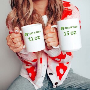 May include: Two white ceramic mugs with green logos and text. The larger mug says "TEES N TEES 15 oz" and the smaller mug says "TEES N TEES 11 oz".
