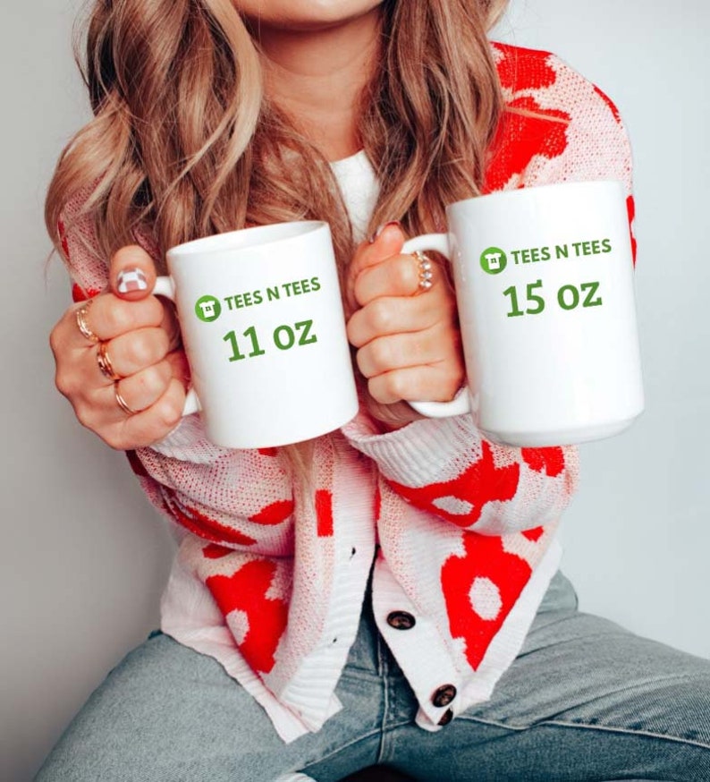 May include: Two white ceramic mugs with green logos and text. The larger mug says "TEES N TEES 15 oz" and the smaller mug says "TEES N TEES 11 oz".