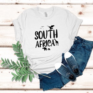 May include: White t-shirt with the words "SOUTH AFRICA" in black, featuring silhouettes of an eagle, birds, giraffe, and elephants. The shirt is paired with blue jeans and navy sneakers with white soles.