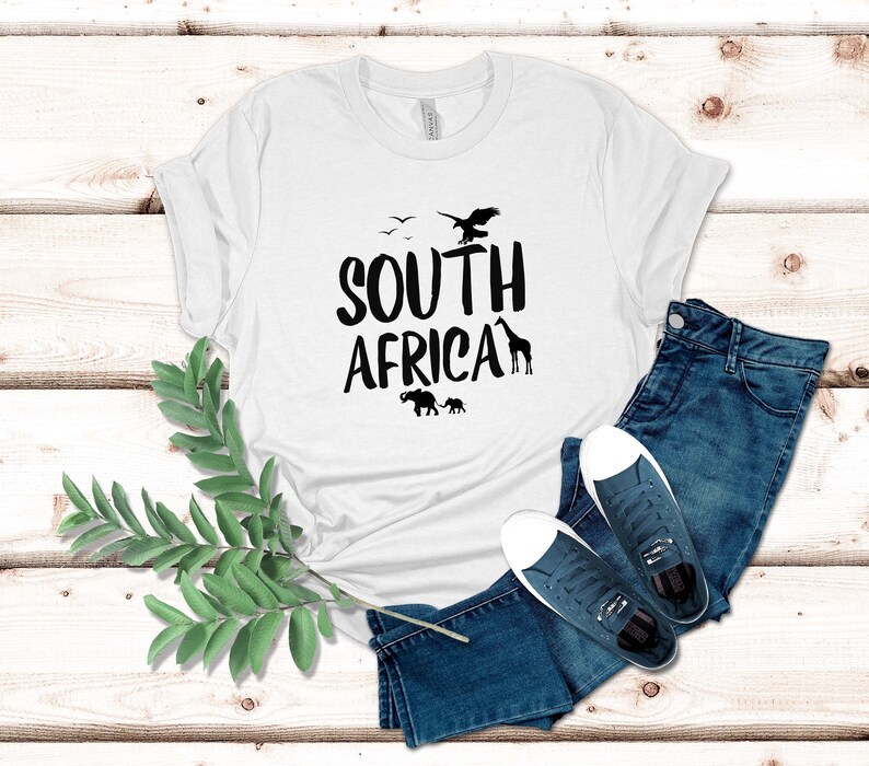 May include: White t-shirt with the words "SOUTH AFRICA" in black, featuring silhouettes of an eagle, birds, giraffe, and elephants. The shirt is paired with blue jeans and navy sneakers with white soles.