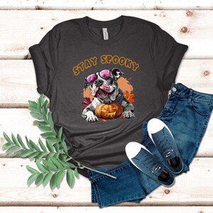 May include: A dark gray t-shirt with the text "Stay Spooky" in orange letters. A cartoon image of a bulldog wearing sunglasses and holding a jack-o-lantern is centered on the shirt.