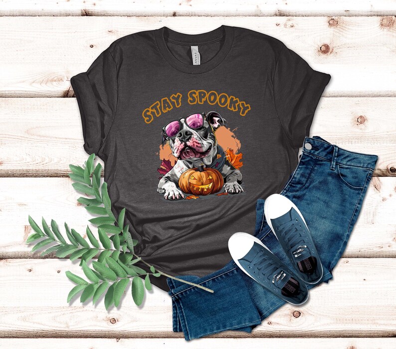 May include: A dark gray t-shirt with the text "Stay Spooky" in orange letters. A cartoon image of a bulldog wearing sunglasses and holding a jack-o-lantern is centered on the shirt.