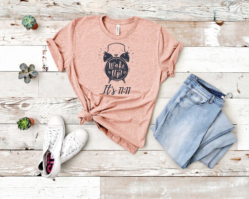 May include: A pink t-shirt with a black alarm clock graphic and the text "Wake Up! It's 11:11". The shirt is laid out on a white wooden surface with a pair of white sneakers and blue jeans.