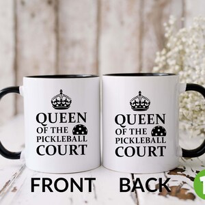 May include: White ceramic coffee mug with a black rim and handle. The mug has a black and white graphic of a crown and the text "Queen of the Pickleball Court" with a pickleball graphic. The mug is shown from the front and back.