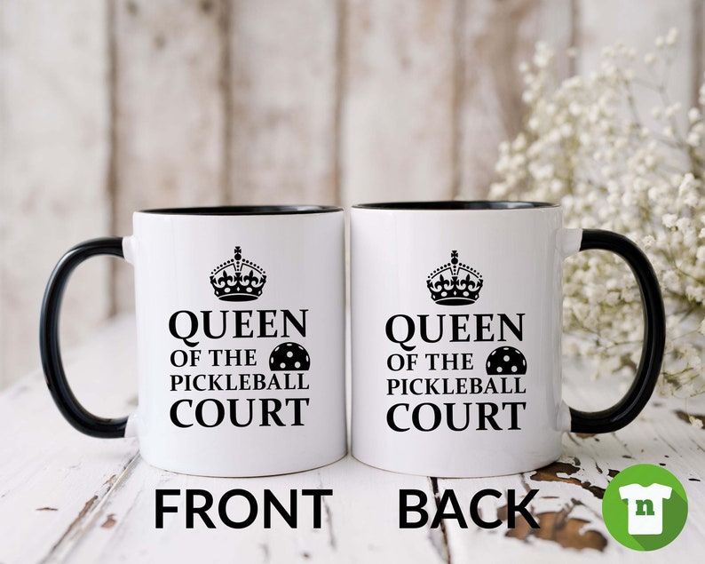 May include: White ceramic coffee mug with a black rim and handle. The mug has a black and white graphic of a crown and the text "Queen of the Pickleball Court" with a pickleball graphic. The mug is shown from the front and back.