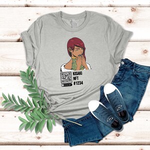 May include: A light gray t-shirt featuring a graphic of a woman with red hair and a QR code. The text "KISAKI NFT #1234" is printed below the graphic. The shirt is paired with blue jeans and dark blue sneakers.