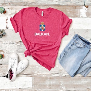 May include: A pink heather t-shirt with a blue and white crest and the word "BALKAN." The shirt is laid out on a white wood background with a pair of white sneakers and a pair of light blue jeans.