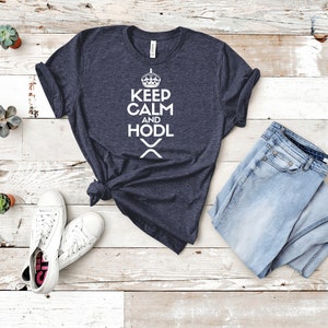 Puede incluir: Una camiseta azul marino con las palabras "KEEP CALM AND HODL" en blanco, un gr&aacute;fico de corona y un s&iacute;mbolo. Se combina con vaqueros azul claro y zapatillas blancas. Peque&ntilde;as suculentas en macetas est&aacute;n dispersas.