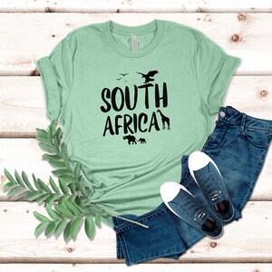 May include: A light green t-shirt with the words "SOUTH AFRICA" in black, featuring silhouettes of birds, a giraffe, and elephants. The shirt is paired with blue jeans and dark blue sneakers with white soles. A sprig of green leaves is in the foreground.
