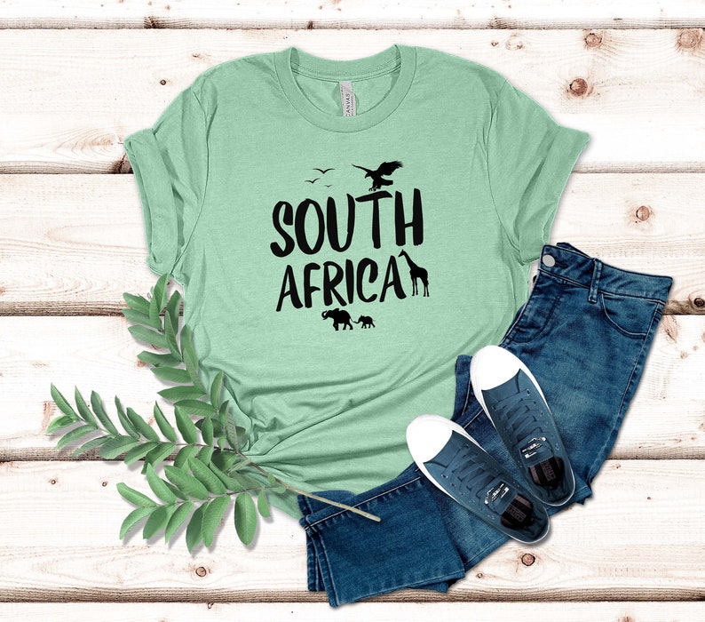 May include: A light green t-shirt with the words "SOUTH AFRICA" in black, featuring silhouettes of birds, a giraffe, and elephants. The shirt is paired with blue jeans and dark blue sneakers with white soles. A sprig of green leaves is in the foreground.