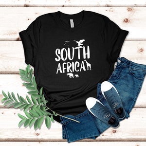 May include: Black t-shirt with the words "SOUTH AFRICA" in white, featuring silhouettes of birds, an eagle, a giraffe, and elephants. The shirt is paired with blue jeans and white-soled sneakers.