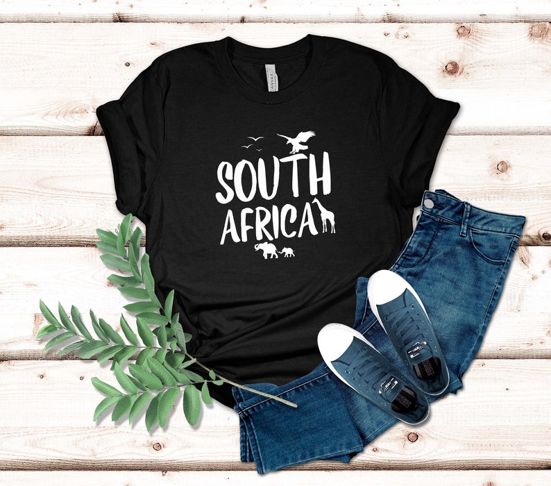 May include: Black t-shirt with the words "SOUTH AFRICA" in white, featuring silhouettes of birds, an eagle, a giraffe, and elephants. The shirt is paired with blue jeans and white-soled sneakers.