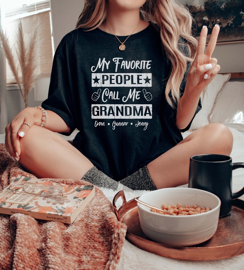 May include: A black t-shirt with the text "My Favorite People Call Me Grandma" and the names "Evan, Gunner, Jenny" printed on it. The shirt is worn by a person sitting on a bed.