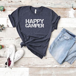 Puede incluir: Una camiseta azul marino con el texto "HAPPY CAMPER" en letras blancas desgastadas. La camiseta est&aacute; colocada sobre un fondo de madera blanca con un par de zapatillas blancas y unos vaqueros azules claros.