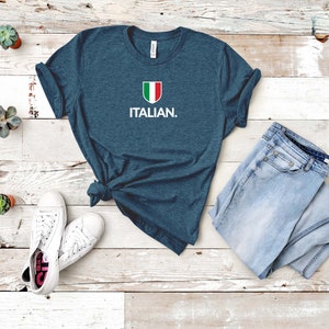 May include: A dark blue t-shirt with a white Italian flag graphic and the text "ITALIAN." The shirt is laid out on a white wooden surface with a pair of white trainers and a pair of light blue jeans.