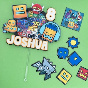 May include: A selection of colourful cake toppers and decorations in a cartoon style. Features a topper with the name "JOSHUA", the number "8", and various geometric and character designs in orange, blue, and yellow. The background is a solid green.