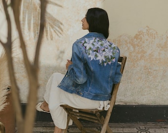 May include: A woman wearing a denim jacket with embroidered flowers on the back. She is sitting on a wooden chair and wearing a white skirt.