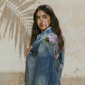 May include: A blue denim jacket with floral embroidery on the back. The embroidery features purple, pink, and green flowers.