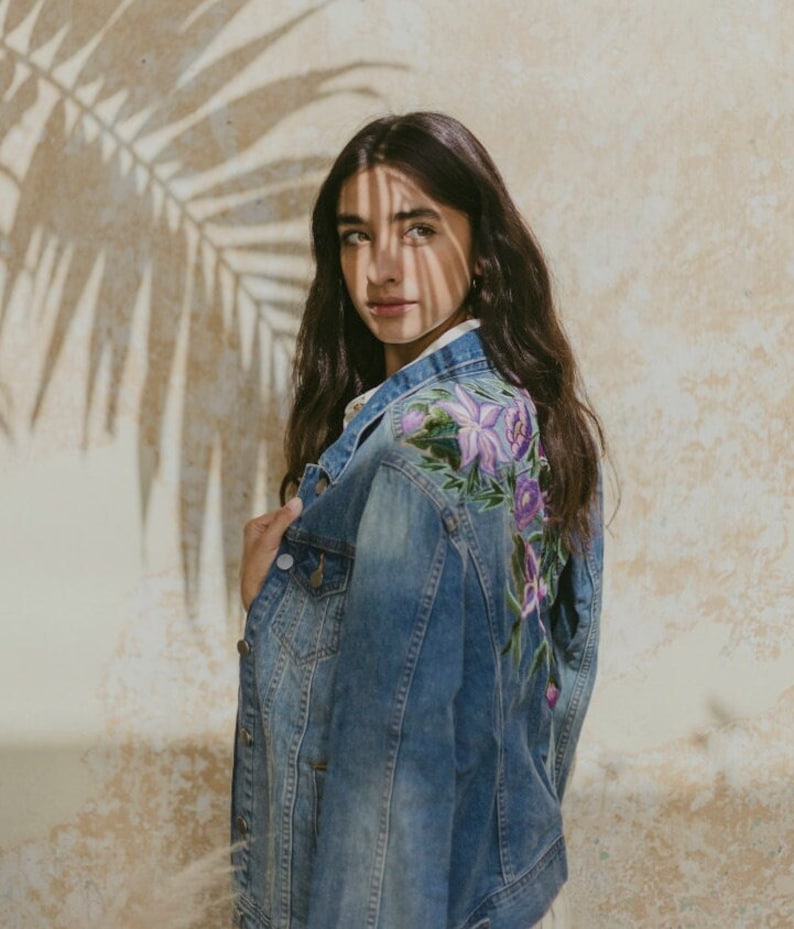 May include: A blue denim jacket with floral embroidery on the back. The embroidery features purple, pink, and green flowers.