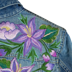May include: A denim jacket featuring embroidered flowers in shades of purple, green, and yellow. The flowers are a mix of lilies and roses.