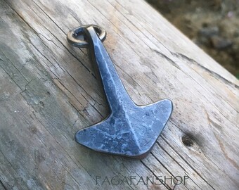 Peut inclure: Un pendentif en métal argenté en forme de marteau avec un anneau en haut. Le pendentif a une surface texturée et repose sur une surface en bois.