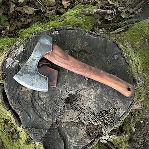 May include: A hand-forged steel axe head with a curved cutting edge attached to a smooth, dark brown wooden handle. A brown leather sheath is tucked beneath the axe head. The text PAGAFANSHOP is visible on the surface of the wood handle.