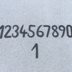 Könnte beinhalten: Schwarze Hausnummern aus Metall von 1 bis 9 und eine zusätzliche 1. Die Zahlen sind in einer Reihe angeordnet, wobei die zusätzliche 1 unter der Reihe liegt.