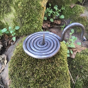 May include: A hand-forged metal candle holder with a spiral base and a central spike. The holder has a curved handle and is set on a moss-covered rock. The candle holder is a unique piece of home decor.