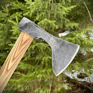 May include: A hand-forged axe with a textured, gray steel blade and a light brown wooden handle. The axe head has a curved shape and a sharp edge. The handle is made of wood with a visible grain. The text "PAGAFANSHOP" is visible on the handle.