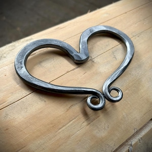 May include: A hand-forged metal heart with a decorative swirl design. The heart is made of iron and has a rustic, aged finish.