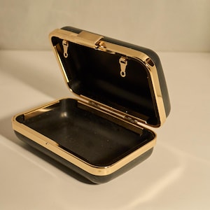 May include: Black rectangular clutch with a gold-colored frame and clasp. The interior is black, and the clutch is open, revealing the inside. The gold trim adds a touch of elegance to this accessory.
