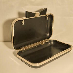 May include: Open black clutch with a gold-colored trim and a bow-shaped clasp. The interior is black, and the clutch is rectangular. The clasp is black with a gold-colored accent.