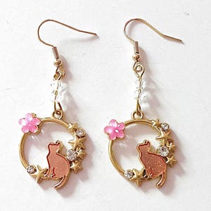 May include: Gold dangle earrings with a pink cat silhouette, pink flowers, and clear crystals. The cat is surrounded by gold stars.