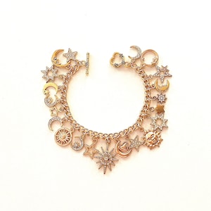 May include: Gold-toned charm bracelet featuring celestial-themed charms. The bracelet includes charms shaped like moons, stars, and suns, some adorned with clear crystals. The chain has a toggle clasp. The bracelet is displayed on a white background.