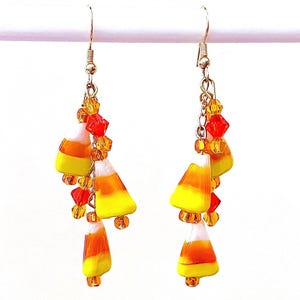 May include: A pair of dangling earrings featuring a candy corn design. Each earring has a silver-colored hook with a chain of orange and yellow beads, and three candy corn charms with yellow, orange, and white stripes.