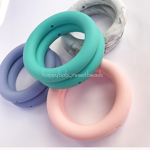 Op de afbeelding: Vier siliconen ringen in verschillende kleuren: lichtblauw, turquoise, roze en wit met een marmerpatroon. De ringen hebben elk twee gaten en zijn op elkaar gestapeld. De tekst "happybabysweetbeads" is zichtbaar op de turquoise ring.
