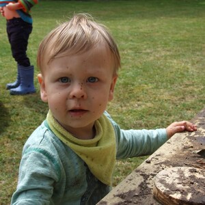 May include: A young boy with blonde hair and blue eyes is wearing a light blue shirt with a yellow bandana. He has dirt on his face and is smiling.