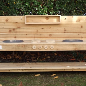 May include: Wooden play kitchen with two sinks, a shelf, and four knobs. The kitchen is made of natural wood and has a rustic look.