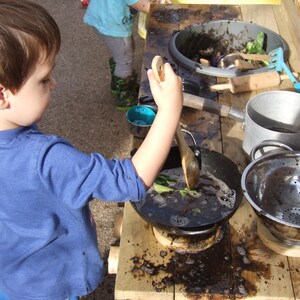 May include: A young child is playing with a toy kitchen set. They are stirring a black substance in a pan with a wooden spoon. There are other toy kitchen utensils and a metal colander on the table.